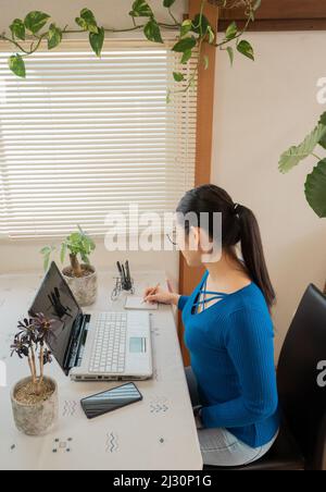 Ragazza asiatica con occhiali, cuffie e piante che lavorano da casa con un computer portatile e cellulare. Foto Stock
