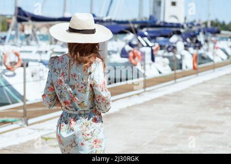 Vista da dietro elegante donna turistica solista in abito floreale con cappello sul molo. Foto Stock