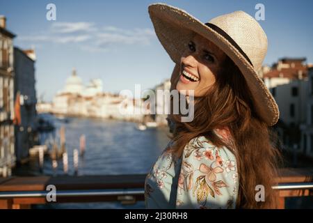 Giovane donna turistica sorridente in abito floreale con cappello che esplora le attrazioni sul ponte dell'Accademia a Venezia. Foto Stock