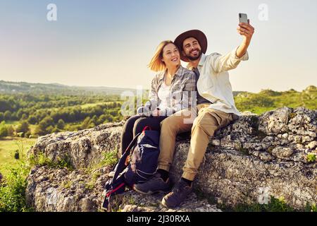 Giovane coppia che prende selfie mentre si siede su roccia in montagna durante l'escursione estiva Foto Stock