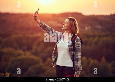 Felice giovane donna che prende selfie durante le escursioni con zaino durante il tramonto Foto Stock