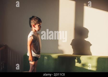Profilo di un ragazzo in piedi sul divano fissando la sua ombra sul muro. Avere un volto carino. In piedi contro l'interno della stanza. Foto Stock