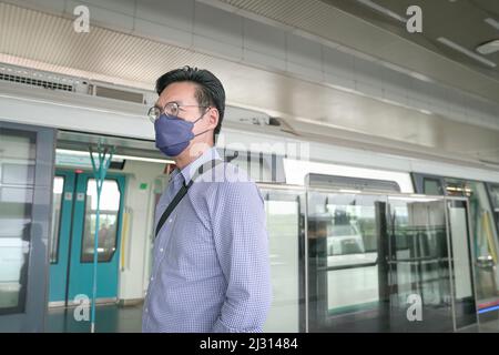 Uomo asiatico commuter con maschera facciale in piedi sulla piattaforma del treno della metropolitana. Concetto di transito mascherato. Foto Stock