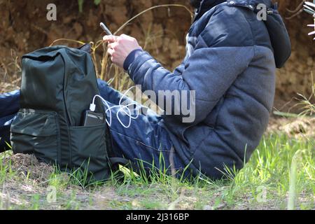 Uomo seduto a terra e tiene uno smartphone nelle sue mani e lo carica con il power bank nero sullo sfondo di una vista della natura. Caricabatterie da viaggio portatile. Foto Stock