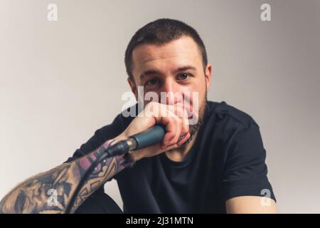 attraente uomo marrone-eyed con un tatuaggio seduto tenendo un microfono in mano e mettendo sulla bocca medio primo piano luce sfondo scatto studio. Foto di alta qualità Foto Stock