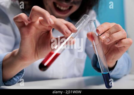 Primo piano di pazzo scienziato goofy mescolando sostanze chimiche liquide per creare una nuova formula mentre si siede alla scrivania. Laboratorio di maniaco che fa siero sperimentale utilizzando composti tossici. Foto studio. Foto Stock