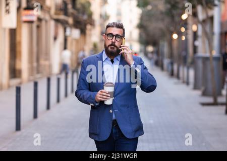 Uomo d'affari che tiene una tazza di caffè e parla al telefono mentre cammina per la strada. Foto Stock