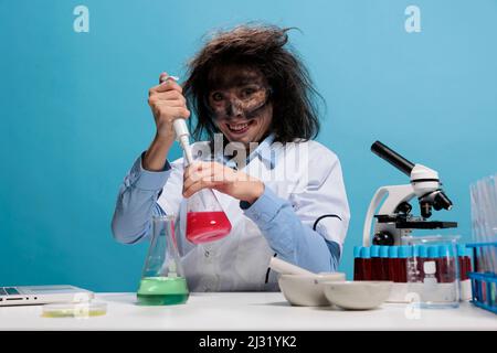Il chimico pazzo che osserva grinning creepy alla macchina fotografica mentre usando il contagocce al lavoro di laboratorio dopo l'esplosione chimica. Stupida lavoratrice di laboratorio con pipetta seduta alla scrivania su sfondo blu. Foto Stock