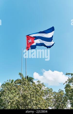 Bandiera nazionale di Cuba ondeggiante in vento su flagpole sopra alberi verdi su sfondo di cielo nuvoloso Foto Stock