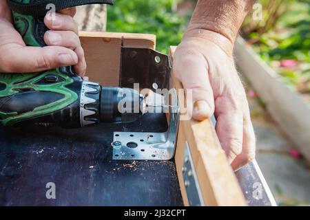 Primo piano. Un carpentiere con le mani protette da guanti trapana una tavola di legno con un trapano elettrico. Riparazioni fai-da-te nella vita reale. Giardinaggio Foto Stock