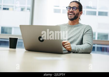Un giovane uomo d'affari allegro ridendo felice mentre lavora con un laptop. Imprenditore di successo che lavora da remoto in uno spazio di co-lavoro. Foto Stock