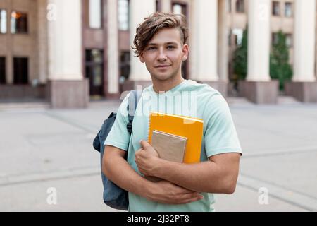 Portraif di ragazzo felice studente con zaino e libri di lavoro che posano su sfondo edificio universitario Foto Stock