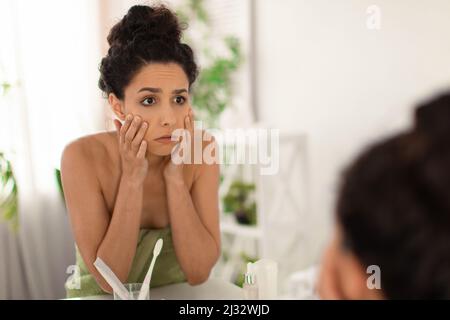 Giovane donna sconvolta che controlla le prime rughe intorno ai suoi occhi, sensazione di stanchezza o stress, guardando a specchio dopo il bagno a casa Foto Stock