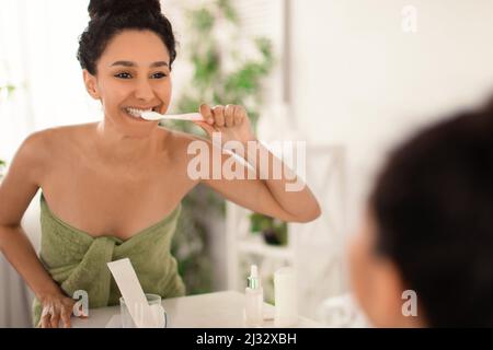 Bella signora giovane con un bel sorriso che indossa un asciugamano dopo il bagno, spazzolando i denti con lo spazzolino vicino allo specchio a casa Foto Stock