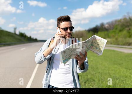 Il turista maschio sollecitato con la mappa in piedi sulla strada, parlando sul telefono cellulare, non può prendere l'auto o trovare il suo percorso di viaggio Foto Stock