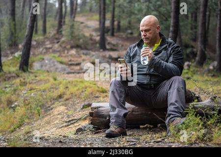 Una persona seduta su un ceppo nei boschi con una bottiglia d'acqua e un telefono in mano Foto Stock