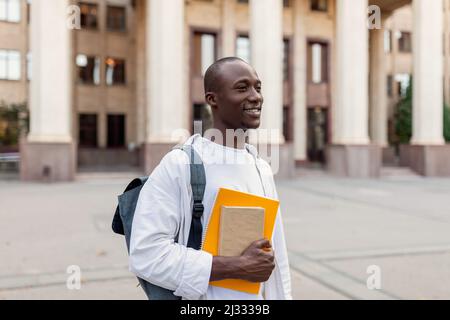 Sorridente studente maschile afroamericano con zaino e libri di lavoro che si posano sul fondo della costruzione del college Foto Stock