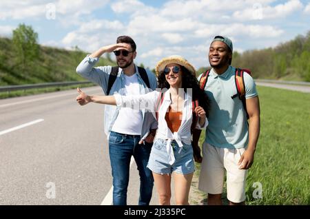 Felice giovani amici multirazziale in piedi in autostrada, mostrando pollice su, hitchhiking per l'auto, andando in viaggio insieme Foto Stock
