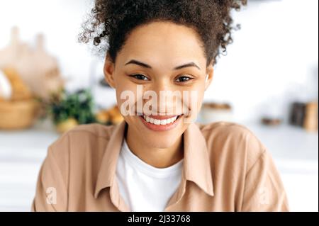Primo piano ritratto di una ragazza allegra e allegra millenaria afroamericana con capelli ricci e frane, con un sorriso bianco neve, indossando una maglietta casual, guardando la macchina fotografica, sorridendo felicemente Foto Stock