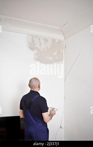 Un janitor o custode di costruzione che ispeziona una macchia d'acqua grande e varie crepe in parete e soffitto del soggiorno di una vecchia casa. Foto Stock