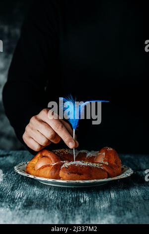 Un uomo sta decorando con una piuma blu una tradizionale torta spagnola mona de pascua, mangiato il lunedì di Pasqua, ornato con uova sode, su un grigio Foto Stock