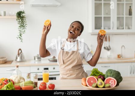 Felice emotivo giovane donna nera in grembiule hanno divertimento su cucina minimalista interno, tiene arance Foto Stock