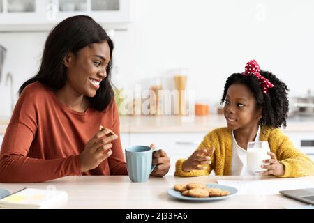 Little Black Girl e la sua mamma mangiando spuntini e chiacchierando in cucina Foto Stock