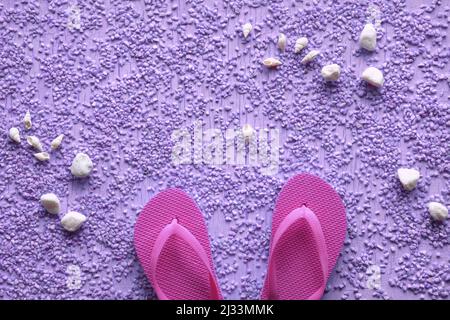 Estate al mare. Infradito in gomma magenta, pantofole su ciottoli con conchiglie e ciottoli, studio concept. Sfondo viola con texture, posto per il testo. Foto Stock