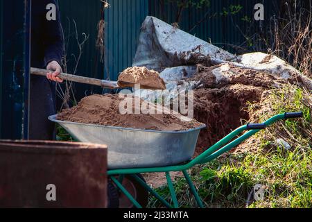 L'uomo usa la pala e riempie la carriola di sabbia. Lavori di costruzione in primo piano. La vita rurale in campagna. Foto Stock