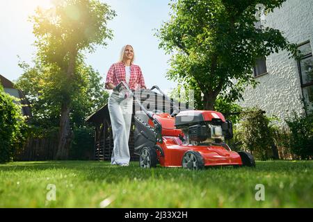 Vista ad angolo basso della bella donna caucasica con capelli biondi utilizzando il rasaerba elettrico per tagliare l'erba sul cortile posteriore. Graziosa donna in abbigliamento casual utilizzando attrezzature moderne per il lavoro in giardino. Foto Stock