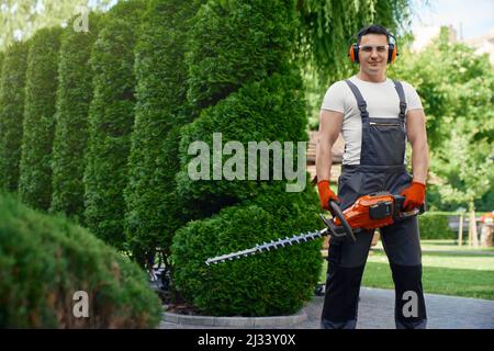 Il giardiniere caucasico competente sorride e guarda la macchina fotografica mentre si trova in piedi sul cortile posteriore e tiene il trimmer elettrico della mano. Uomo che usa l'attrezzo di giardinaggio per tagliare la siepe. Foto Stock