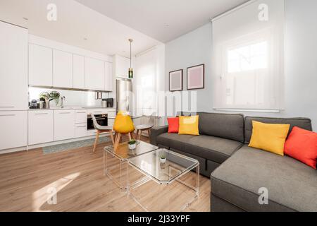 Cucina bianca aperta al soggiorno con tavoli da caffè in metacrilato e divano a tre posti in tessuto grigio con chaise longue Foto Stock