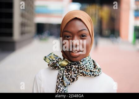 Affascinante femmina afro-americana musulmana in hijab tradizionale ed elegante sciarpa legata al collo in piedi sulla strada della città e fissando alla macchina fotografica Foto Stock