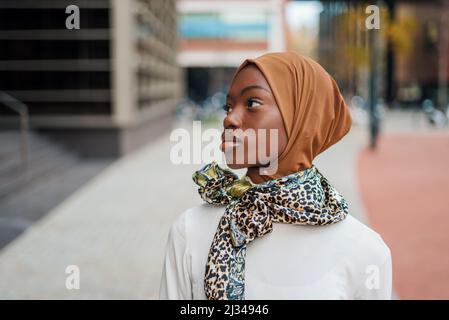 Curiosa femmina islamica afroamericana in foulard tradizionale guardando a lato di un alto edificio nel moderno quartiere cittadino Foto Stock