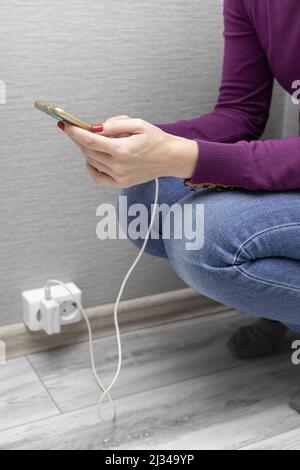 donna che squattava tenendo il telefono collegato al caricabatterie Foto Stock