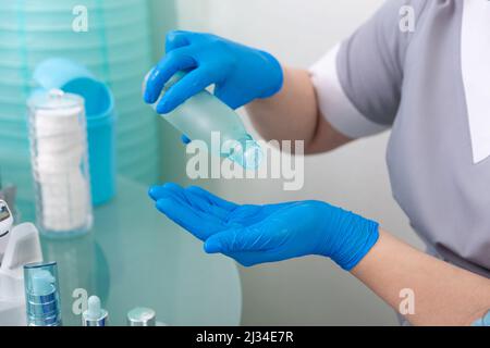 Cosmetologist in maschera medica e cappotto grigio versa toner di levigatura sulla sua mano in guanti blu monouso. Foto Stock