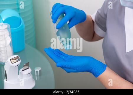 Cosmetologist in maschera medica e cappotto grigio versa toner di levigatura sulla sua mano in guanti blu monouso. Foto Stock