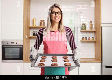 Giovane casalinga con un grembiule che tiene un vassoio di biscotti cotti in una cucina Foto Stock