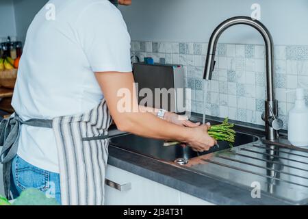 Donna lavaggio fresco asparagi mazzo sotto acqua corrente su lavandino sulla sua cucina. Preparazione degli ingredienti alimentari per il pranzo. Cucinare cibo sano. Ricette Foto Stock