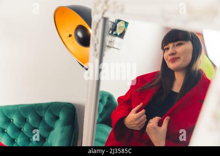 Giovane donna caucasica dai capelli scuri che prova su cappotto rosso. Guardando lo specchio ritratto copia spazio . Foto di alta qualità Foto Stock