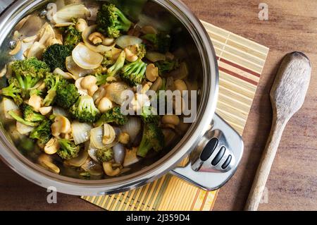 Padella con broccoli saltati, funghi, aglio affettato e cipolle tritate grossolanamente, la padella si trova su piccoli tappeti di bambù con un cucchiaio di legno pla Foto Stock