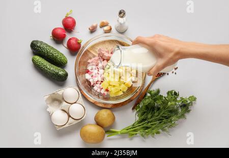 Donna che versa il kefir in una ciotola con ingredienti per l'okroshka su sfondo grigio Foto Stock