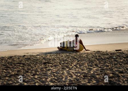 Surfista seduto sulla spiaggia di sabbia guardando il mare. Spiaggia di Farol da barra a Salvador, stato di Bahia, B. Foto Stock