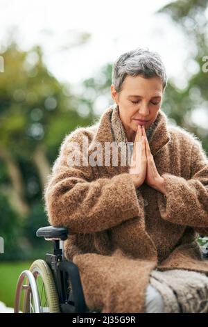 Ricerca di guida dall'alto. Scatto corto di una donna anziana che prega mentre si siede in sedia a rotelle all'aperto. Foto Stock