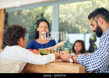 Dacci oggi il nostro pane quotidiano. Shot di una famiglia che prega insieme prima di gustare un pasto al tavolo. Foto Stock