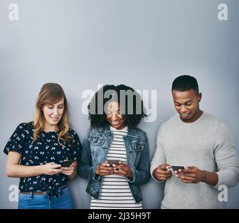 Connessione alla stessa rete wireless. Studio girato di tre giovani utilizzando i loro cellulari su uno sfondo grigio. Foto Stock