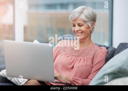 Sembra davvero sorprendente. Scatto di una donna matura allegra usando il suo laptop mentre si rilassa sul suo divano a casa. Foto Stock