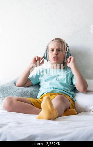 I bambini che indossano le cuffie ascoltano la musica. Ragazzo che ascolta la musica in cuffia seduto sul letto. Foto Stock