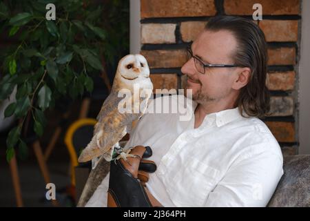 Uomo con gufo a casa. Animali domestici insoliti e rapporti umani di amicizia animale. Uccello selvatico a portata di mano guanto tenerezza e amore. Foto Stock