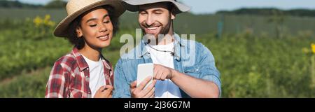 giovane agricoltore che mostra smartphone al allegro collega afroamericano, banner Foto Stock